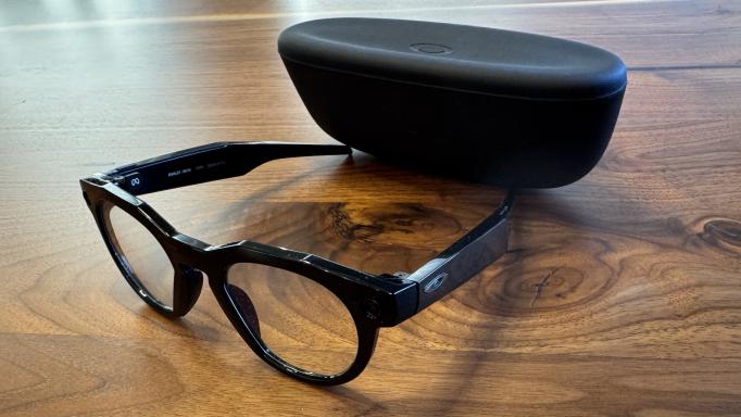 Photo of black framed Oakley Meta Glasses with the case which is matte black.  Both are sitting on a dark wood table.